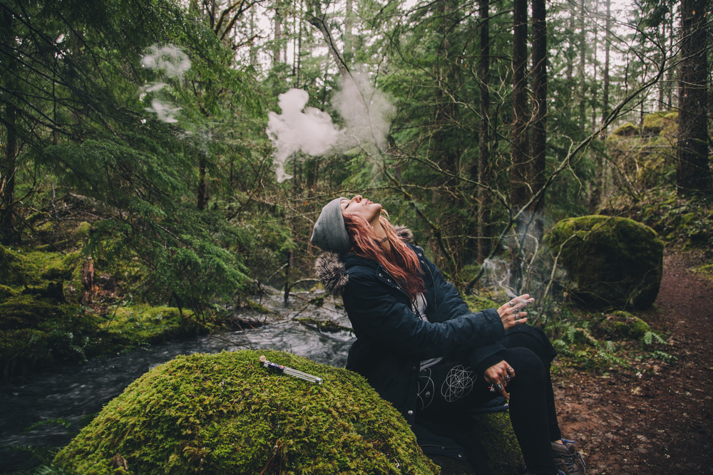 A woman takes a smoke break in a mossy forest from her vape pen.